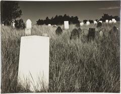 A black and white photograph of a grave with others in the background, positioned on a plot of tall grass. Trees appear in the distance.