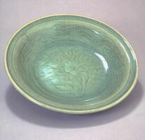 This stoneware, flat-bottomed dish has straight, everted sides and an everted rim with articulation, on a footring. The interior is incised with a peony spray surrounded by wavy lines on the sides. It is covered with a green celadon glaze.
