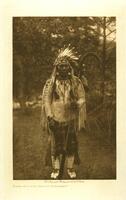 This is a photograph of a man standing outdoors, grass and trees surrounding him. He holds a large, curved staff in his left hand. He wears decorated clothing and a feather headdress. 