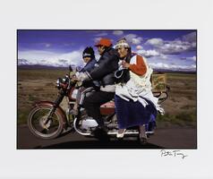 An image of a boy, man, and woman atop a maroon motorbike which appears to be in motion. They travel on a paved road through a plain. 