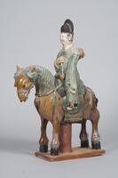 An earthenware polychrome glazed figure of a horse and rider.  The amber-colored horse has a green mane and black taill and is standing on all fours on a platform.  It has large eyes and a full saddle and reigns.  The male rider is dressed in long green robes with long sleeves, a tall black hat, and the face is painted in mineral pigments. 