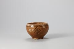 <p>This cup is similar in appearance to a round pot, with its mouth curving inwards slightly. The glaze has been oxidized, producing brown tints. The outer rim is black inlaid with a fret-patterned band, below which is also inlaid in black with chrysanthemum spray designs in three places. Coarse sand is stuck on the entire foot and outer base. Glaze has owed down to the interior of the cup during ring and has severely peeled off from the mouth and the outer surface in parts, exposing the body.<br />
[<em>Korean Collection, University of Michigan Museum of Art</em> (2014) p.123]<br />
&nbsp;</p>
