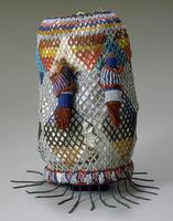 Cylindrical headdress with diamond-shaped multi-colored beadwork over a cloth base. The bottom rim has a pattern of red and blue triangles outlined on the edge with red. A fringe of blue beads hangs all the way around the bottom edge. There are two beadwork birds attached to the front of the headdress. 