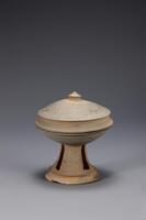 Flared base with rounded food storage bowl on top. The base is cut with evenly spaced rectangular holes. The lid is incised with a repeating herringbone, or dotted design. The know on the lid is the shape of a Buddhist canopy, or chattra.<br />
<br />
This is a dark blue-gray, high-fired stoneware lidded stem cup. The lid is crowned by a pearl-shaped knob, while both the inner and outer surfaces of lid have traces related to the attachment of the knob to the lid. A v-shaped pattern of engraved dots, made using a sixtooth comb, surrounds the central knob. The cup&rsquo;s flange slopes inwards and has a sharp edge. The cup body has a horizontal gallery that holds the lid in place. The stem is perforated by rectangular openings, below which is a sharply protruding circular raised band. Traces of rotation and water smoothing are visible on the body and stem of the cup.
<p>[Korean Collection, University of Michigan Museum of Art (2017) p. 59]</p>
