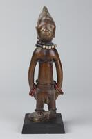 Standing human figure with hands at the sides mounted on a square base. One of the feet and part of the original base have broken off. There are strings of beads around the waist, wrists, and neck as well as brass around the ankles. On the face there are incised grooves and the hair is in the shape of three triangular lobes decorated with horizontal lines. 