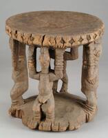 Wooden stool with a circular base and four human figures supporting the upper portion. The figures have horizontal grooves decorating their wrists and ankles. The edge of the upper portion of the stool is decorated with incised diamond shapes. 