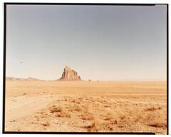 This photograph depicts a desert landscape with a craggy rock formation rising in the far distance in the central portion of the horizon. A dirt road extends through the foreground. The top half of the image depicts a vast, cloudless sky in tones of blue and pink.