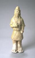 An earthenware standing figure of a military official or warrior, wearing armor that includes a helmet, elbow-length gauntlets, a cuirass with plaques, and taces, worn over a long tunic, loose pants, and boots.  The arm is raised to hold a weapon. The top half is covered in a straw-colored glaze.  One of a pair with 1997/2.25.