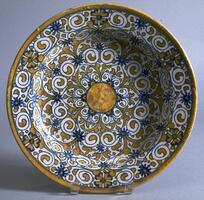 This maiolica dish is covered by a radial pattern consisting of abstract leaves and curling tendrils punctuated with flowers all centered around a yellow disk in the center of the well. The harmony of the design stems from radial symmetry of the pattern as well as the restricted palette of blue and a soft brass-yellow.