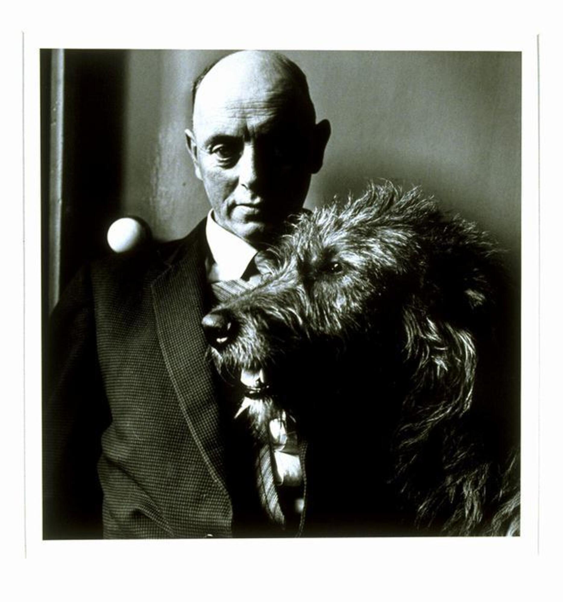 This photograph depicts a middle-aged man in a suit seated with his dog. The two figures sit inside a domestic space.