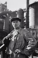 A man in worker's blues and hat looks off toward the right-hand side of the photograph as he holds a cigarette in his right hand.