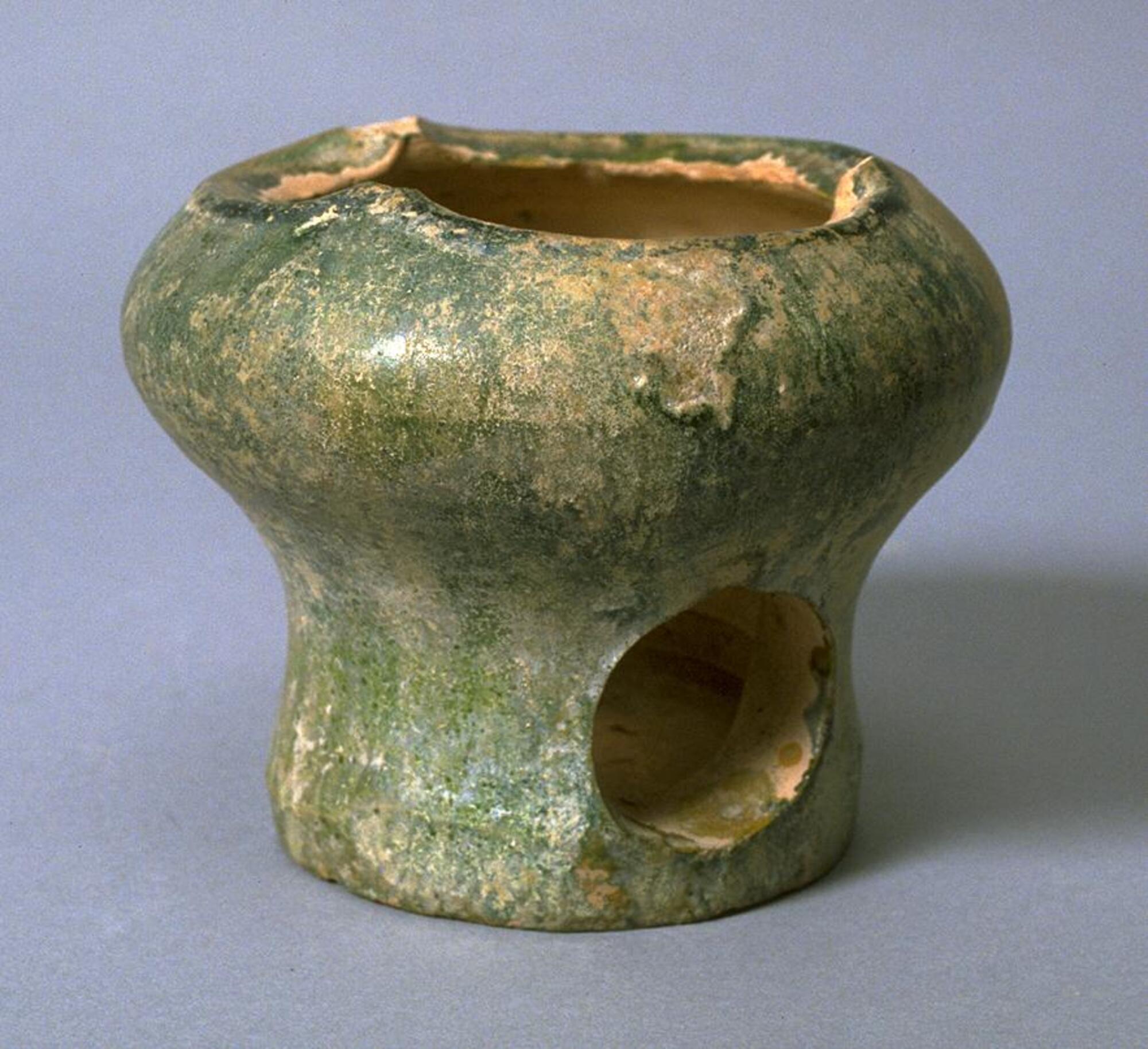 A red earthenware, small, flat-bottomed pot with narrow base and wide bulbous neck with three protrusions at the rim, a large circular hole pierced into the side of the lower body, covered in a green lead glaze with iridescence and calcification. 