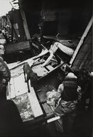 A man wrapped in stained cloth lays on a fallen interior door. Three men and a dog surround him on the wall of a house that has been torn from its frame. A headboard and mattress are buried in other construction debris just behind the group of men.