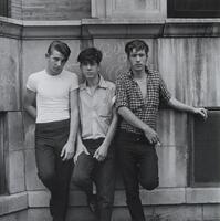 A photograph of three young men standing in front of an outdoor, concrete wall. The man on the left wears a white T-shirt, the man in the middle a button-down, and the man on the right, an unbuttoned shirt, holding a cigarette in his right hand.