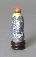 Blue and white snuff bottle with painted mountain landscape scene with a house, trees, hills and mountains. It has green tinted ivory collar and a coral stopper.
