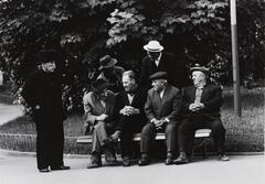 Image of a group of seven men sitting or standing around a bench. One man stands slightly off to the side, hands in pockets, dressed in all black