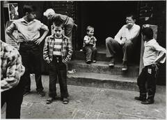 Seven children play on and around the cement stoop of a brick building. One stands with arms akimbo, while another looks towards the pavement with hands in pockets.