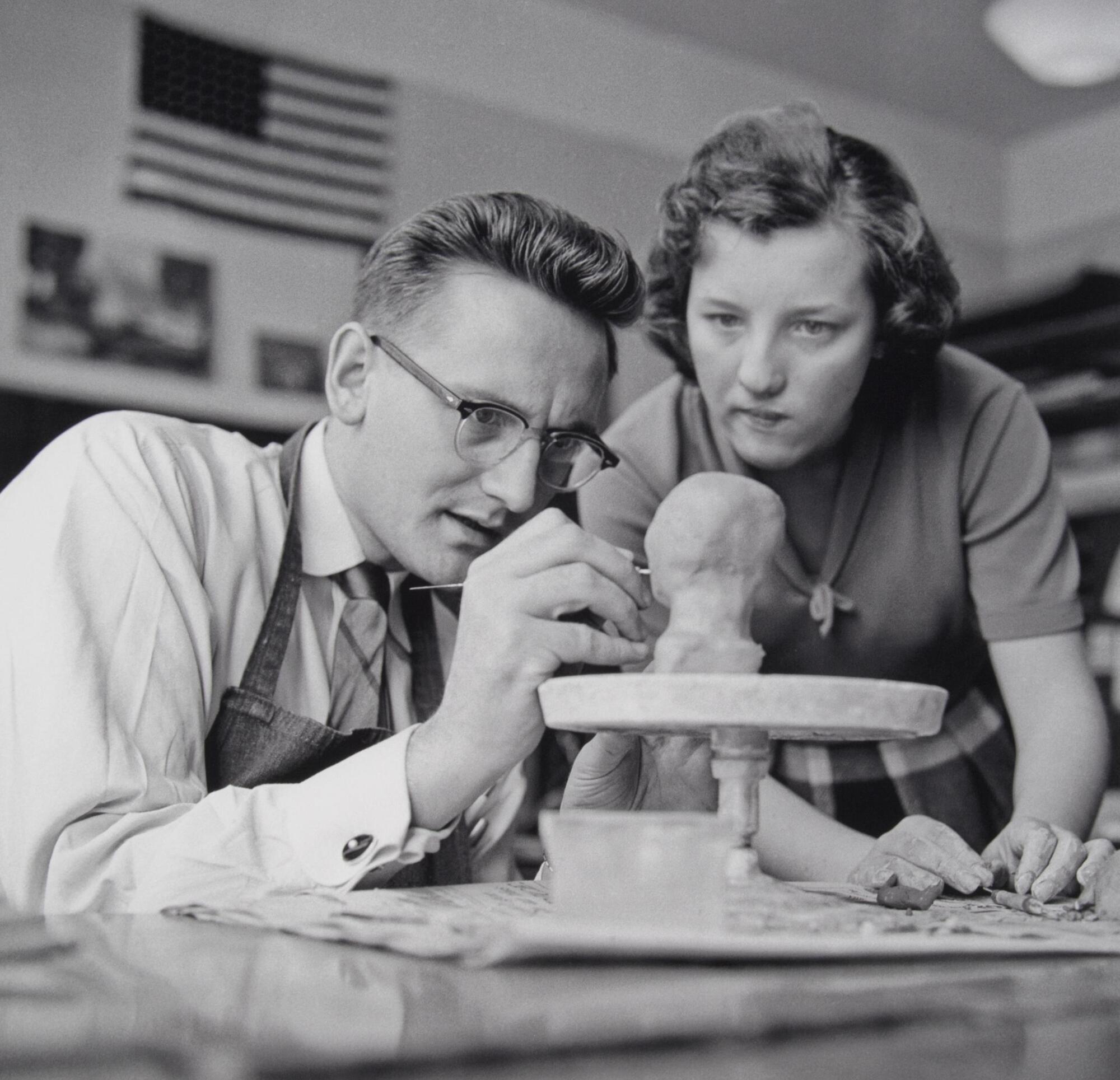 A man and woman working on a sculpture project that is seated on a lazy susan. The man is holding a small metal tool and the woman is looking over his shoulder.