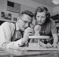 A man and woman working on a sculpture project that is seated on a lazy susan. The man is holding a small metal tool and the woman is looking over his shoulder.
