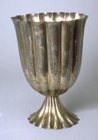 This vase consists of two fluted elements: the container which has a slightly flaring lip, and a foot that is also fluted. The surfaces of the vase have a hand-worked character and are not smoothly finished