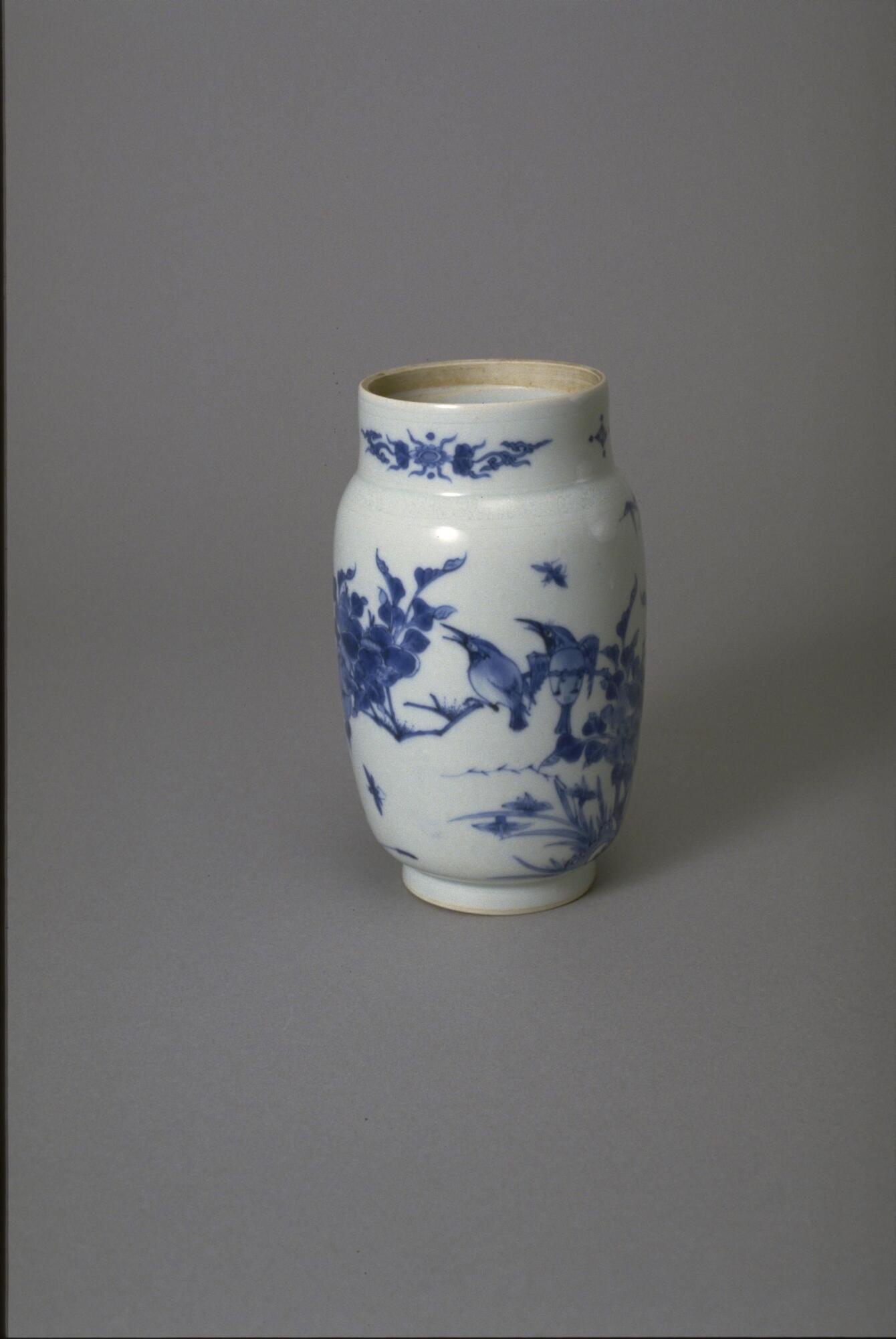 This porcelain jar with cylindrical body, has a recessed neck and foot with the same diameter. The body is decorated with underglaze blue paintings of magpies, plum branches, and bamboo, with floral scrolls around neck. The jar is covered with a clear glaze and is missing the dome capped lid. 