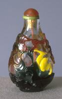 A glass snuff bottle with red and green colored grounds and a yellow bat. On the top is a carnelian stopper in a green tinted ivory collar.