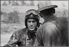 A photograph of two men. The man in the foreground has his back to the camera, conversing with the second man, who wears a jean jacket, helmet, motorcycle goggles, and faces toward the camera.