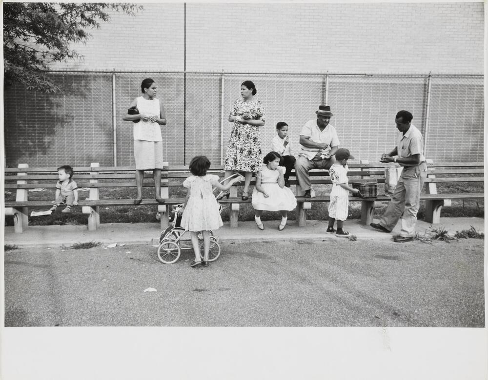 Two women, two men, and five children gather at a row of benches in front of a chain link fence and brick building.