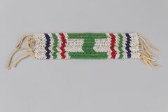 Beaded jewelry with a rectangular piece made up of white beads with green "H" shape in the middle and three stripes on either end of blue, green and red beads. Twisted fringe at two ends.