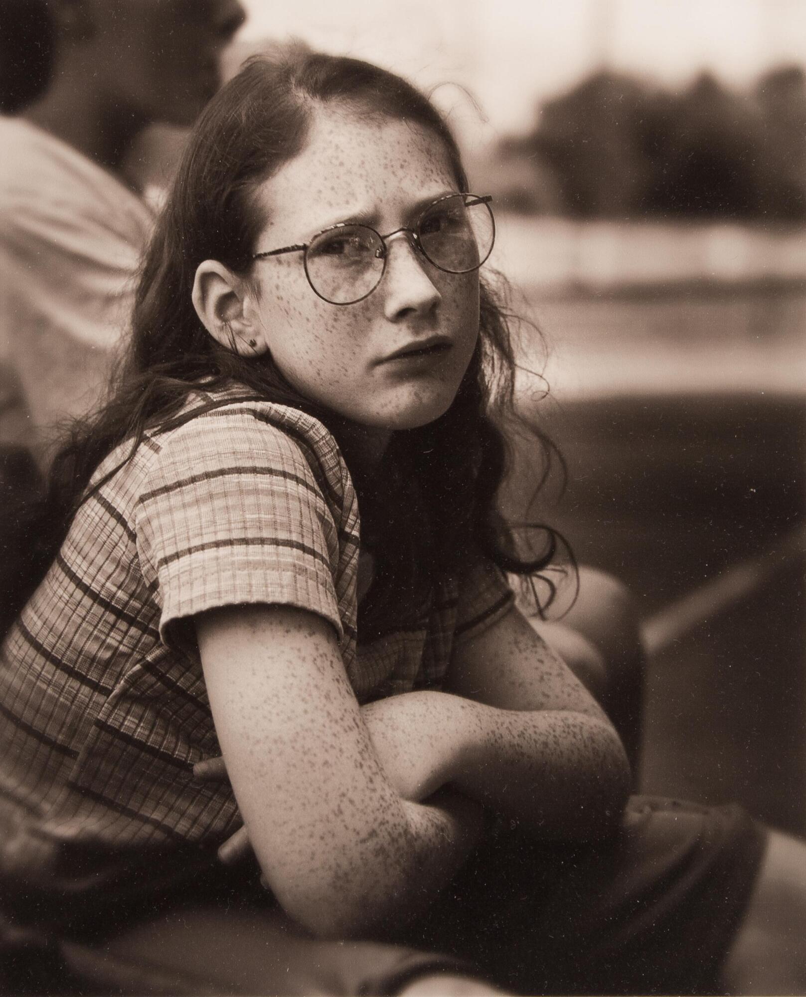 Girl with freckles, arms crossed, wearing glasses.