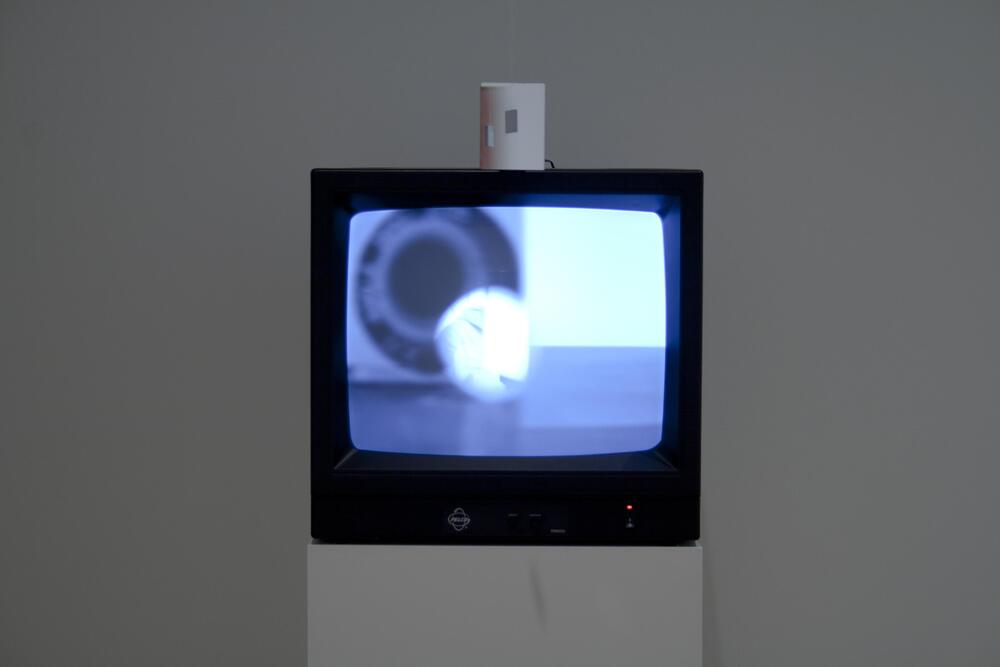 An installation piece that consists of a television monitor on a stand with a closed-circuit camera mounted on top. The lens is pointed directly off the top of the monitor, but there are mirrors suspended from the ceiling that change the camera's perspective.