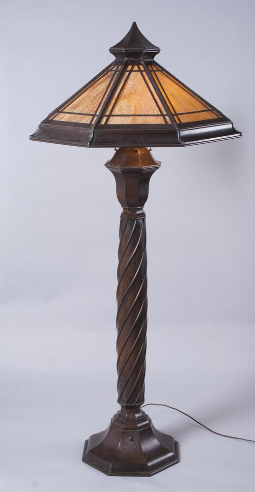 The peaked shade has eight panels with overlaid linear copper border decoration above a cove molded lower rim. The lamp retains four original sockets. The upper column consists of an octagonal capped vase-form with a dominant ogee profile over a round medial column which is slightly beveled in form and decorated with spaced, spirally arranged brass rods applied to its surface. The octagonal molded base repeats the ogee profile of the upper vase form.