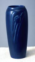 Slender blue-glazed vase with a streamlined form that tapers to a base narrower than the shoulder of vessel. The work has a modeled and stylized motif of peacock feathers. Overall effect is of simplicity and refinement.