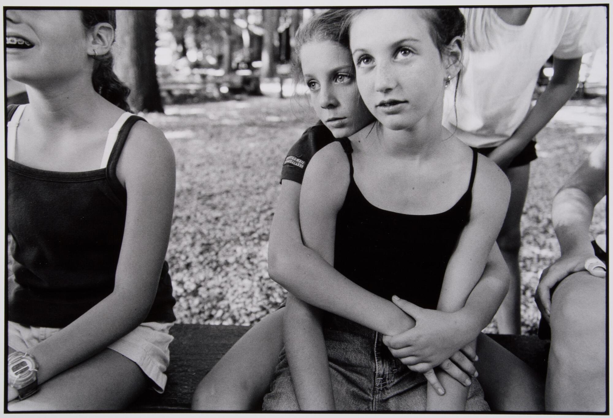 Three girls are seated on a bench and one girl is behind the other giving her a hug. The girls in the front are wearing dark tanktops. The girl in the back is wearing a darker tshirt.