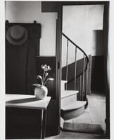 Photograph of the interior of a room, with a flower in a vase on a table, a hat perched on a hook on the wall behind it, and, through a doorway, a view of the bottom steps of a curved staircase.