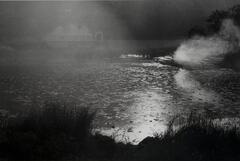 A black and white photo of fog over a pond. A bridge can be seen through the fog.