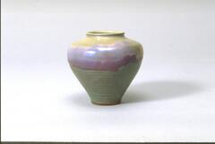 This vessel has a thin mouth that widens to a broad shoulder before tapering to a comparatively slender base. The colors of the glaze include a rich mustard yellow near the mouth, transitions to iridescent shades of purple o the shoulder and then a deep celadon green on the lower portion of the vessel.