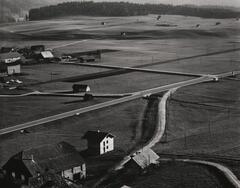 A black and white aerial view of farms and their surrounding land. A forest can be seen in the background.