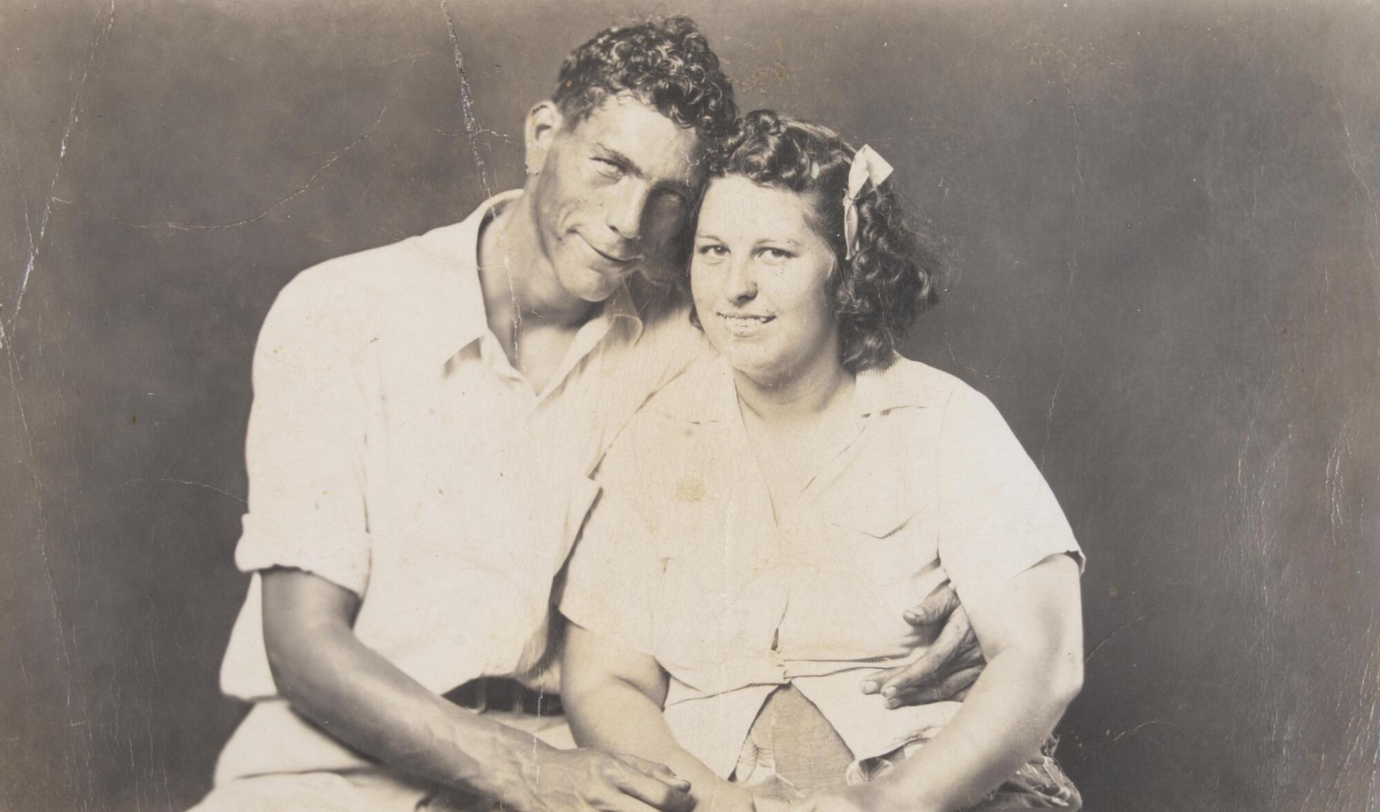 A portrait of a seated couple. A man sits on the left, his arm around a woman to the right. She wears a ribbon in her hair. Both wear light-colored, short-sleeved shirts.