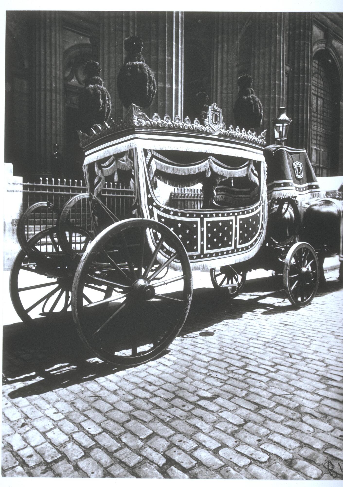 Horse-drawn funeral carriage parked on the side of a cobble-stone paved street.