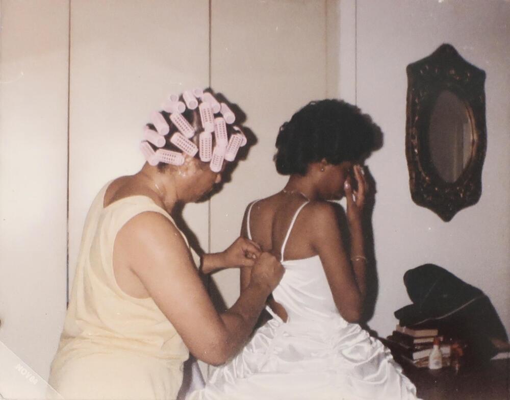 A woman with pink curlers in her hair adjusts the back of a young woman&#39;s dress. Both of them are facing away from the camera and the young woman has her right hand raised to her face.