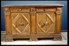 The front of this handsome oak credenza, or sideboard, is divided into halves by three pilasters. Each half is outfitted with a drawer and a door below. The decorative and functional components are artfully arranged across the front of the piece to form a balanced composition of repeated geometric shapes and harmonious proportions.