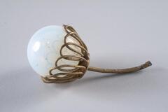 <br />
A clouded glass orb in a brass-colored metallic cap comprised of looped petal-like shapes connected to a thin metal stem.
