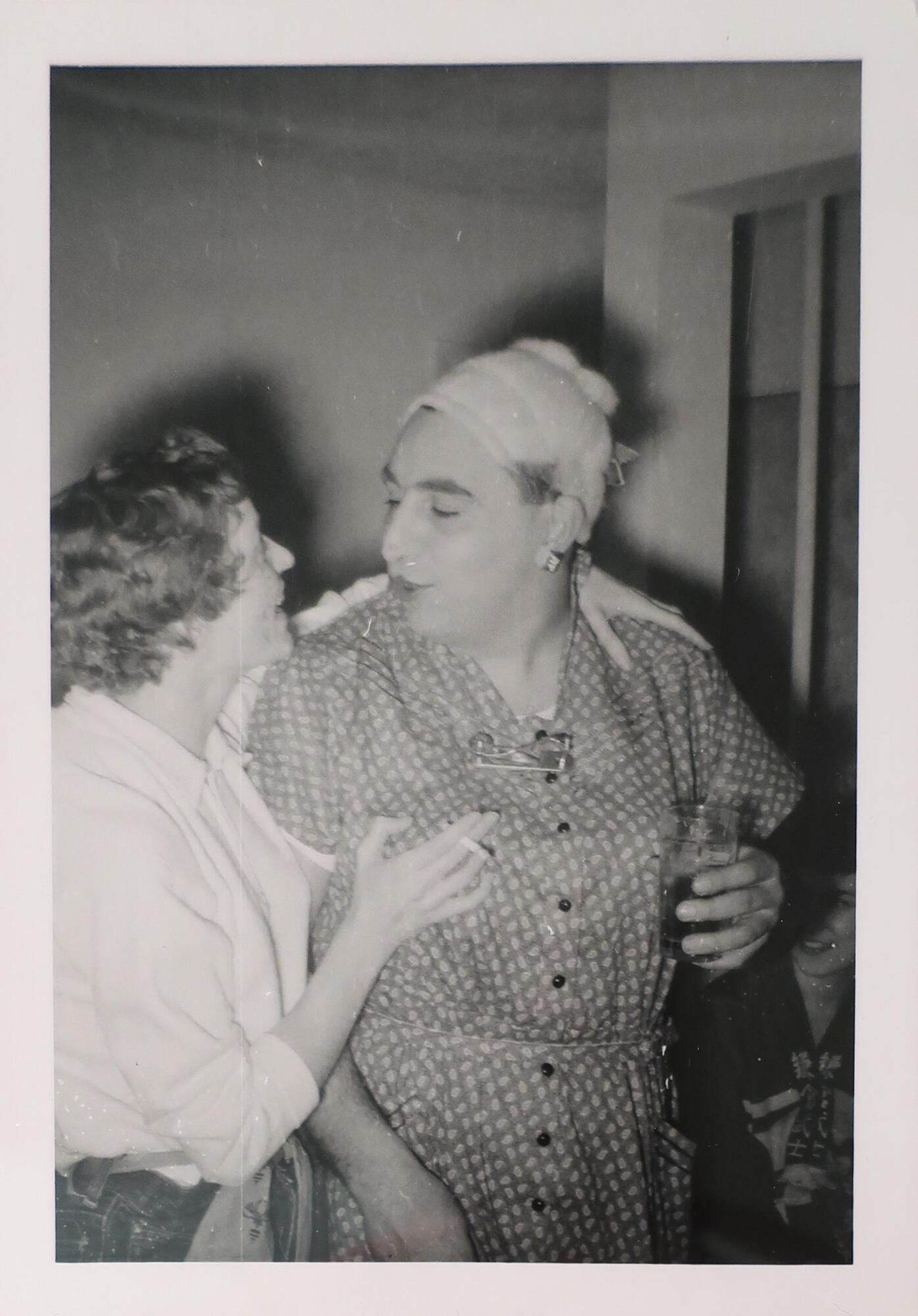 Two people talking to each other, one wearing a print dress with their hair wrapped and the other with a short haircut and white collared shirt. The person in the collared shirt has their left arm around the person in the dress and holds a cigarette in their right hand.