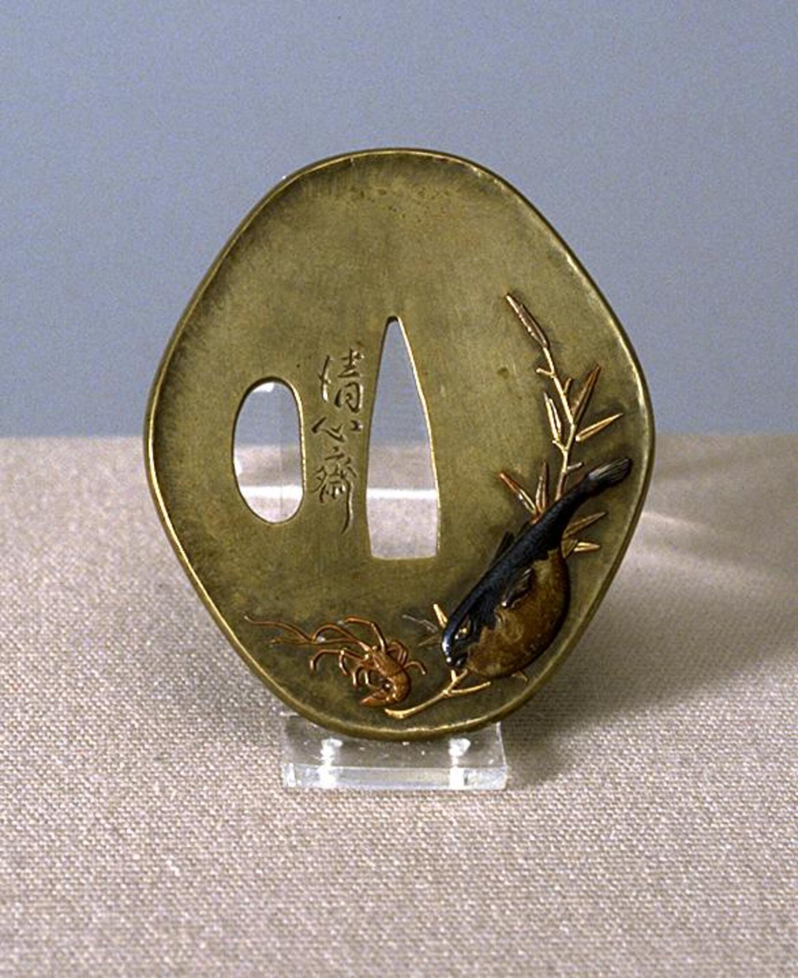 This small, flat piece made of light brown brass (called "sentoku" in Japanese) has a round diamond shape. It has a triangular shaped hole in the center and another round hole on one side. Artist’s name is signed between the two holes. The surface is slightly concaved from the rim. The front has relief design of a shrimp, blowfish, and bamboo branch. On the back, there are designs of a spiral shell, a barnacle, and water drops. The sea motifs are inlayed with gold, silver, copper, and shakudô (copper-gold alloy).