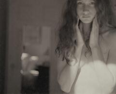 This photograph is of an unclothed young woman holding her hands to the sides of her face. Light from a window illuminates her stomach.