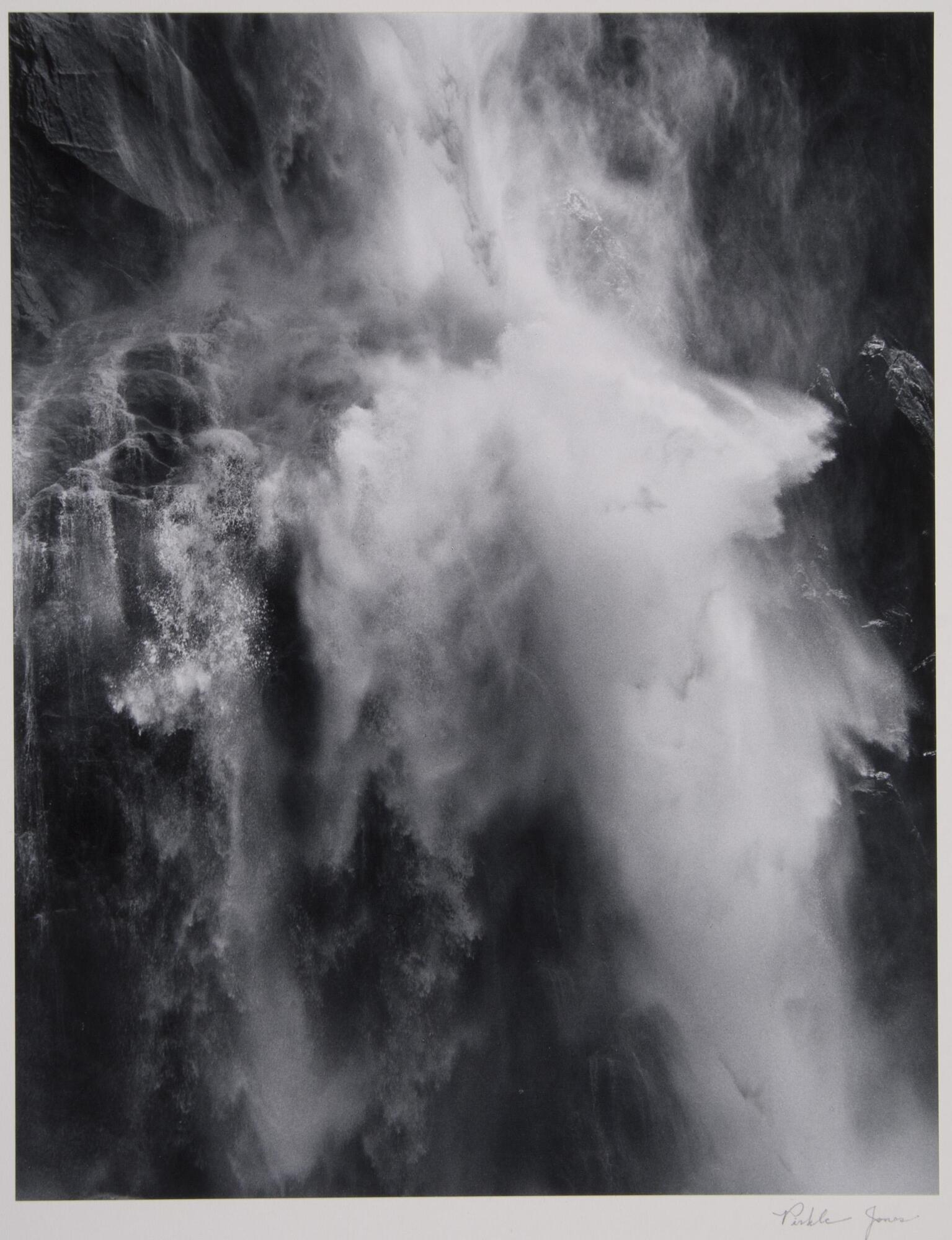 This vertical image is of a waterfall flowing down the side of a hill. It is the middle of the waterfall and the upper and lower portions are not visible. The water on the right hand side appears to be stronger than the left, creating a larger splash.