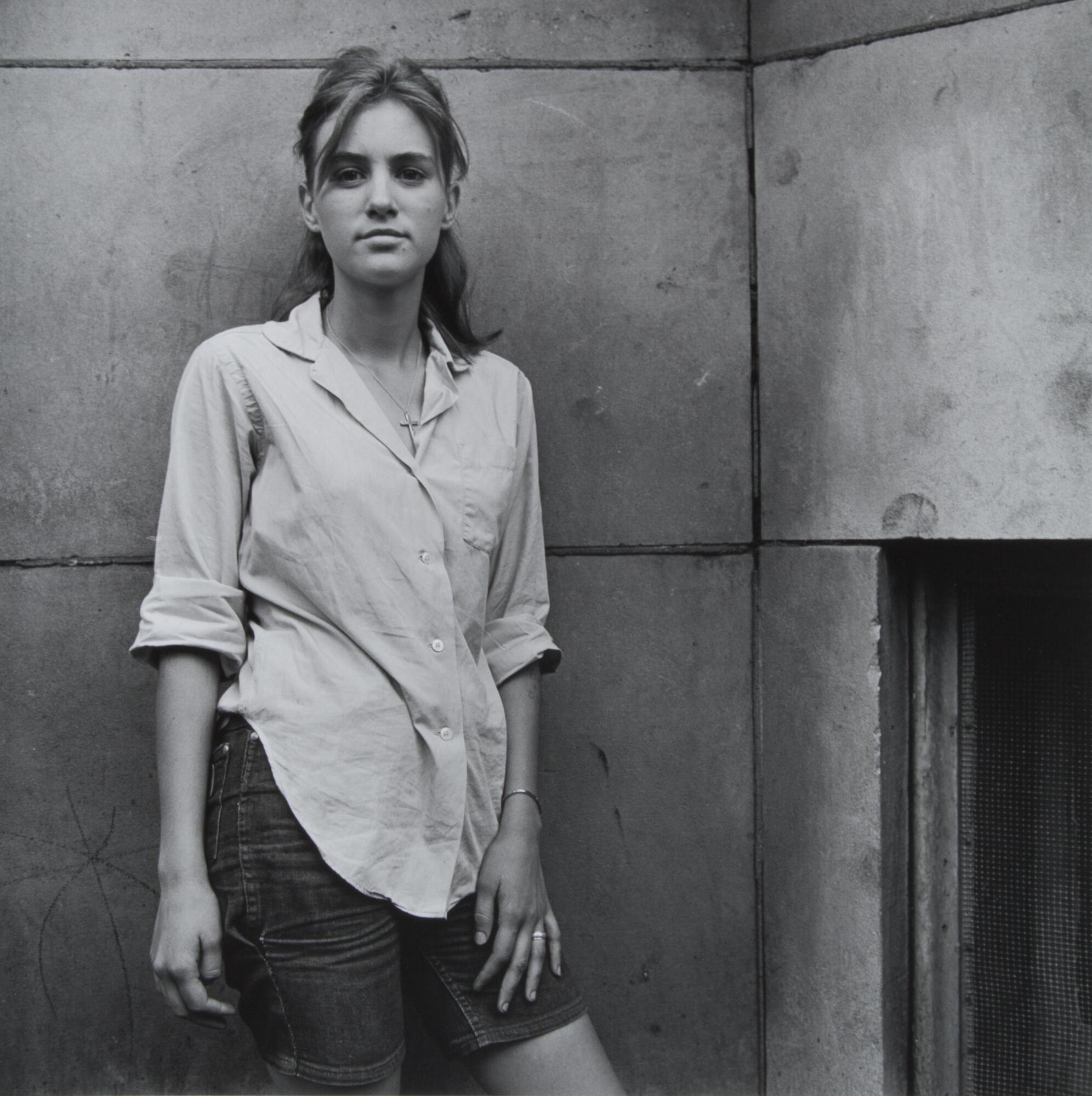 Portrait of a young woman leaning against a concrete, exterior wall. She wears a cross necklace and a button-down shirt that drapes over her mid-length shorts. She looks directly into the camera lens.