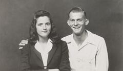 A bust-length portrait of a couple. On the left, a woman in a dark jacket and collared blouse poses with her arms crossed, smiling at the camera. The man on the right wears a light-collared shirt and smiles widely with his arm around the woman.