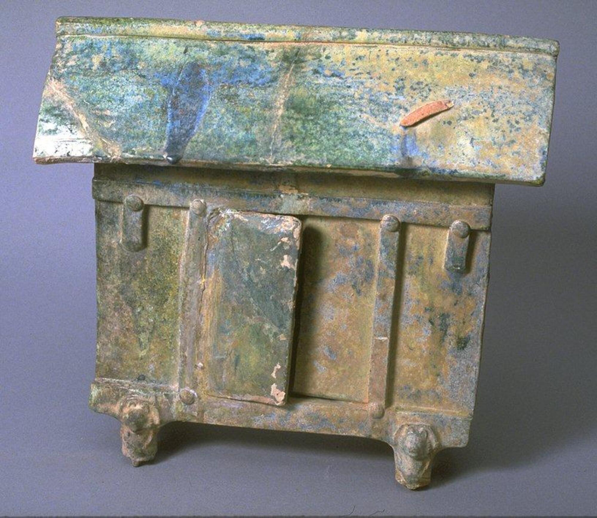 A red, four-sided rectangular earthenware structure in the form of a three-bay storehouse with a peaked roof and ridgeline. Post and lintel details from wooden architecture, with double open doors, on four bear-shaped stilts. The model is covered with a green lead glaze, with iridescence and calcification. 